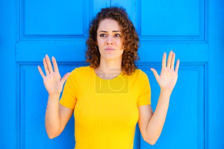 Téléchargez les photos : Jeune femme effrayée en t-shirt jaune avec les cheveux bruns bouclés regardant loin avec les mains levées sur fond bleu sur la rue de la ville - en image libre de droit