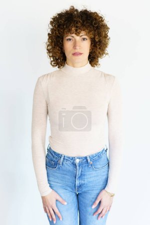 Téléchargez les photos : Femme adulte en col roulé beige et jean aux cheveux bouclés marron regardant la caméra tout en restant immobile avec les bras suspendus droit sur les côtés - en image libre de droit