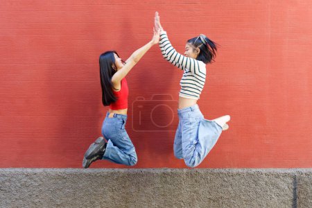 Foto de Cuerpo completo vista lateral de alegre joven asiática mujeres amigos saltando y dando alta cinco el uno al otro contra la pared roja - Imagen libre de derechos