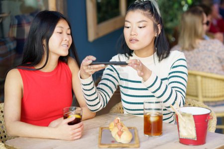 Photo for Smiling young Asian girl friends, in casual clothes sitting at table with cold drinks and taking picture of tasty food on smartphone while spending time together in cafe - Royalty Free Image