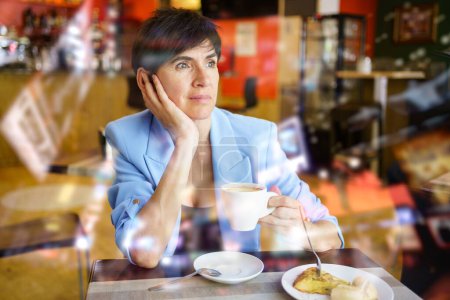 Foto de Mujer meditando en elegante chaqueta azul sentado en la mesa con taza de bebida caliente y mirando hacia otro lado durante el desayuno en la cafetería - Imagen libre de derechos