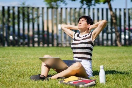 Photo for Full body of peaceful middle aged female freelancer, in summer clothes sitting on grass with laptop and closed eyes while stretching neck during remote work on project - Royalty Free Image