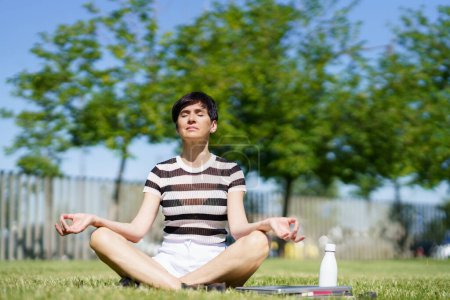 Foto de Cuerpo completo de hembra adulta relajada en camiseta a rayas, sentada en césped cubierto de hierba en pose de loto con los ojos cerrados y las manos de Gyan mudra mientras medita durante la sesión de yoga - Imagen libre de derechos