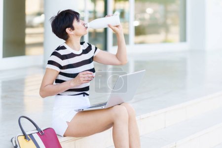 Photo for Concentrated adult female freelancer in casual summer outfit sitting on stairs and drinking water from bottle while working remotely on laptop on street - Royalty Free Image