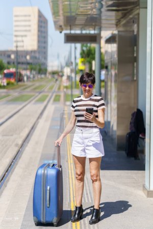 Photo for Full body of cheerful young female traveler in casual clothes and sunglasses smiling and messaging via smartphone while standing with suitcase during summer trip - Royalty Free Image