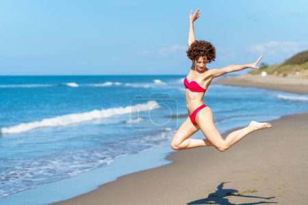 Téléchargez les photos : Vue latérale de la jeune femme en forme auto-assurée, avec les cheveux bruns bouclés en bikini rouge sautant avec les bras levés et regardant vers le bas sur la plage de sable près de la mer ondulée - en image libre de droit