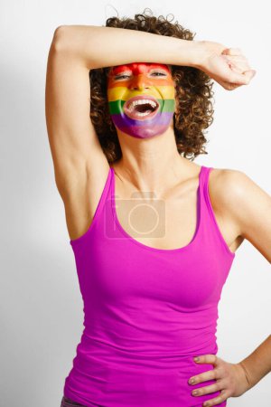 Photo for Cheerful female wearing purple tank top standing with hand on waist and painted in rainbow colors face looking at camera against white background - Royalty Free Image