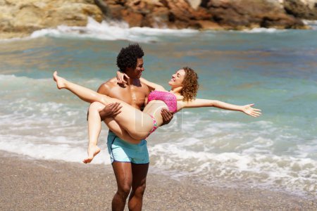 Téléchargez les photos : Heureux homme afro-américain élevant femme avec les mains tendues en maillots de bain tout en se regardant pendant les vacances contre la mer agitant - en image libre de droit