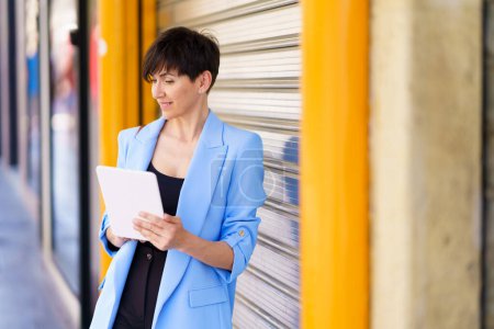 Photo for Positive female entrepreneur in formal clothes standing by silver rolling door with yellow column on street while browsing tablet in daylight - Royalty Free Image