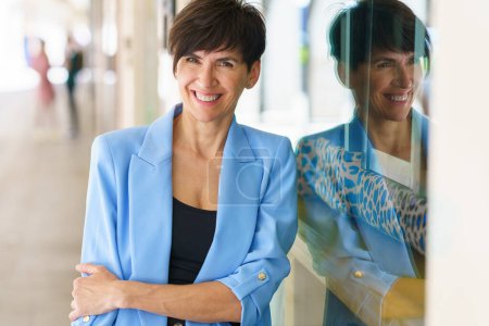 Téléchargez les photos : Femme positive d'âge moyen en veste bleue aux cheveux bruns courts regardant la caméra et souriant tout en s'appuyant sur le mur de verre du bâtiment moderne - en image libre de droit