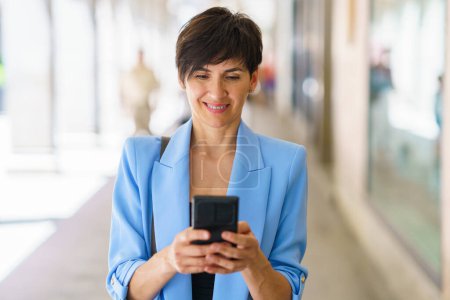 Foto de Alegre mujer de mediana edad en chaqueta azul sonriendo y mensajería en el teléfono móvil mientras está de pie en la calle soleada durante el paseo - Imagen libre de derechos