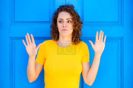 Photo for Afraid young female in yellow t shirt with curly brown hair and raised hands looking away against blue wall on street - Royalty Free Image