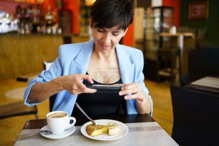 Photo for Smiling young female in blue jacket sitting at table, with cup of coffee and looking at screen of mobile phone while taking picture of snack in restaurant - Royalty Free Image
