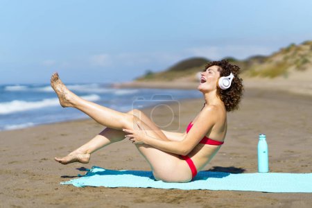 Téléchargez les photos : Vue latérale d'une jeune femme joyeuse en bikini et avec des cheveux bouclés écoutant de la musique avec des écouteurs assis avec la jambe levée sur le rivage - en image libre de droit