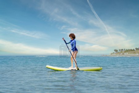 Téléchargez les photos : Surfeuse concentrée en combinaison bleue debout sur le SUP board et pagayant en mer contre un ciel bleu nuageux lors d'une journée d'été ensoleillée - en image libre de droit