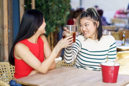 Téléchargez les photos : Délicieuses jeunes amies asiatiques, en tenues décontractées souriantes et se regardant tout en buvant des verres à table en bois dans un café pendant le week-end ensemble - en image libre de droit