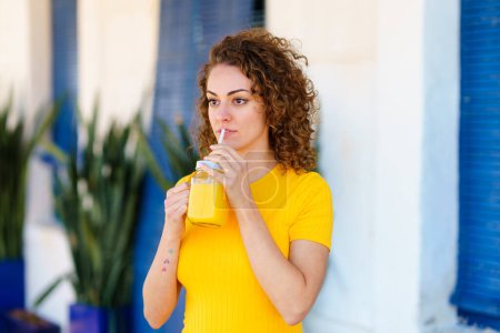 Photo for Pensive young female in yellow t shirt with curly brown hair looking away and drinking fresh juice from glass jar with straw on blurred street - Royalty Free Image
