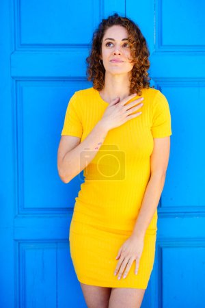 Photo for Dreamy young female in yellow dress with curly brown hair keeping hand on chest and looking up against blue wall on street - Royalty Free Image