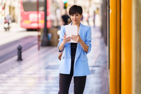 Photo for Concentrated female entrepreneur in formal clothes, reading on tablet while standing on city street and looking at screen against blurred background of people and bus in daylight - Royalty Free Image