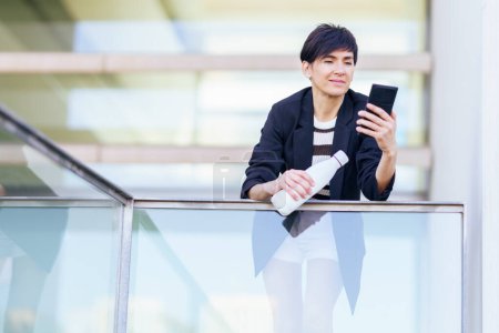 Photo for Smiling female in black jacket holding hand bottle while surfing internet on mobile phone and leaning against glass wall of building with transparent wall - Royalty Free Image