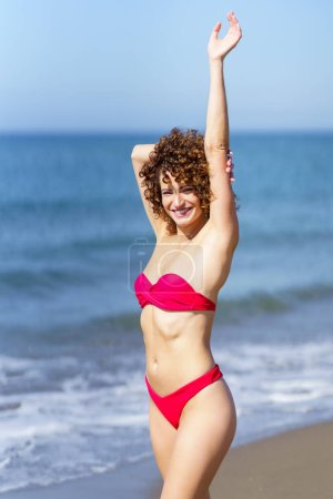 Photo for Cheerful young female redhead in pink bikini standing and touching outstretched arm with hand from behind head, while looking at camera in sunlight against blue seawater and sky - Royalty Free Image