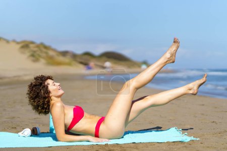 Téléchargez les photos : Vue latérale du gingembre heureux femelle en bikini rose couché sur la plage avec les jambes levées sous un soleil éclatant - en image libre de droit