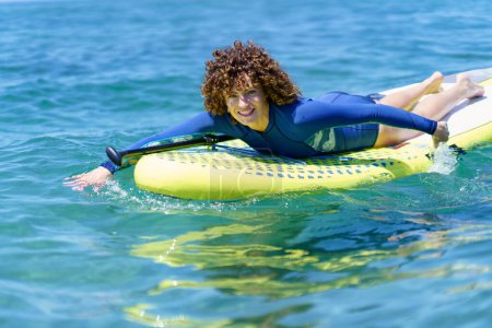 Téléchargez les photos : Heureuse femme bouclée en combinaison de plongée regardant la caméra tout en étant allongée et en tirant la planche à pagaie à travers la surface brillante de la mer au soleil - en image libre de droit
