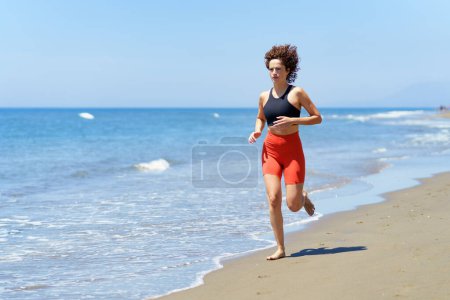 Téléchargez les photos : Jeune femme pieds nus concentrée en vêtements de sport et aux cheveux bouclés regardant ailleurs, tout en faisant du jogging en plein jour sur une plage de sable près de l'eau de mer mousseuse contre un ciel bleu flou - en image libre de droit