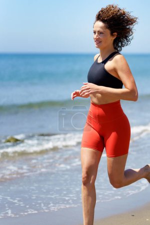 Téléchargez les photos : Vue latérale de la jeune femme pieds nus en vêtements de sport avec les cheveux bouclés souriant et regardant loin, tout en faisant du jogging en plein jour sur la plage de sable près de l'eau de mer mousseuse contre le ciel bleu flou - en image libre de droit