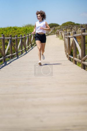 Foto de Cuerpo completo de atleta femenina en forma en ropa deportiva corriendo por el camino de madera y mirando hacia otro lado durante el soleado día de verano - Imagen libre de derechos