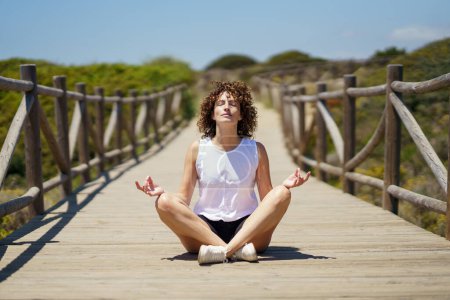 Photo for Young curly haired female in casual clothes sitting on wooden walkway and meditating, with closed eyes while doing lotus pose against blurred green hill under blue sky - Royalty Free Image