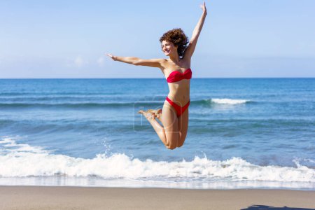 Foto de Mujer joven excitada en bikini rosa que estira los brazos y salta en el aire con las piernas dobladas hacia arriba, mientras sonríe y aleja la vista a la luz del día contra el mar ondulante y el cielo azul - Imagen libre de derechos