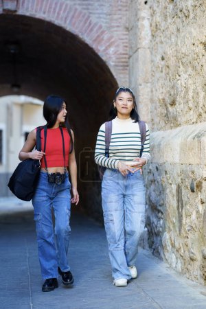 Téléchargez les photos : Corps complet de jeunes femmes asiatiques touristes en vêtements décontractés avec des sacs et un appareil photo se promenant contre un mur de pierre tout en parlant les uns aux autres - en image libre de droit