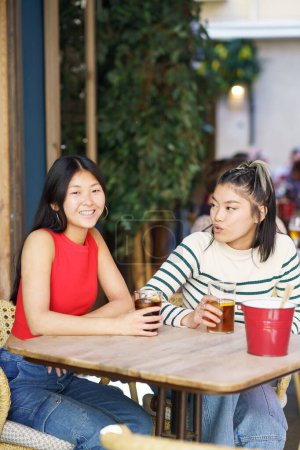 Téléchargez les photos : Joyeux jeunes amies asiatiques, en vêtements décontractés assis à table avec des boissons froides et parlant de plans pour l'avenir tout en passant du temps ensemble dans un café - en image libre de droit