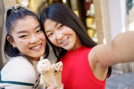 Photo for Positive young Asian women smiling happily and taking selfie while eating delicious ice cream cone together in street - Royalty Free Image