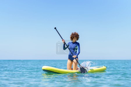 Téléchargez les photos : Focalisé femme bouclée cheveux en combinaison avec pagaie à genoux sur SUP conseil tout en pagayant sur la mer bleue sous ciel bleu sans nuages - en image libre de droit