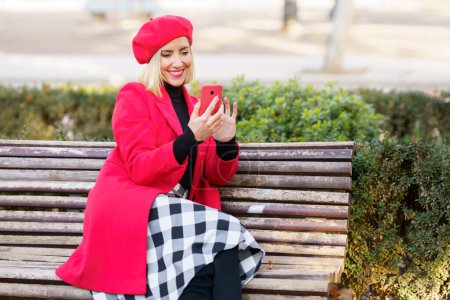 Téléchargez les photos : Joyeuse femme en tenue tendance naviguant sur un téléphone portable moderne assis sur un banc près de plantes vertes dans le parc le jour de l'automne - en image libre de droit