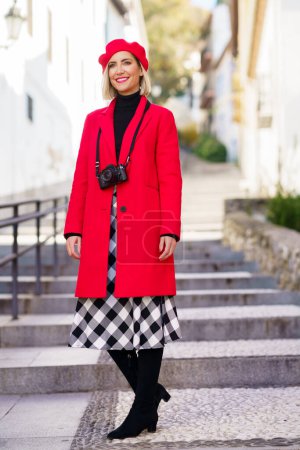 Photo for Full length of happy adult female tourist, in elegant coat and beret with photo camera on neck smiling and looking away while standing on stone stair in old town - Royalty Free Image