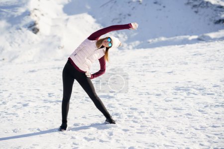 Photo for Full body of young female in sportswear and eyeglasses, stretching arm overhead to one side while practicing yoga exercise on snowy slope in winter at daylight - Royalty Free Image