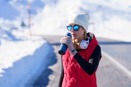 Photo for Athletic woman in outerwear and sunglasses drinking from thermos bottle having workout on road in winter mountains - Royalty Free Image