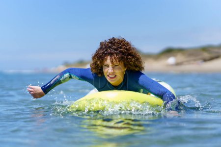 Téléchargez les photos : Jeune surfeuse aux cheveux bouclés en combinaison de plongée allongée sur un SUP board et pagayant les mains flottant au-dessus de la mer contre le ciel bleu - en image libre de droit
