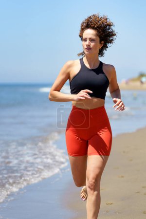 Téléchargez les photos : Souriant cheveux bouclés pieds nus jeune femme en tenue de sport échauffement et détournement des yeux, tout en faisant du jogging seul sur la plage de sable près de la mer agitant contre ciel bleu flou - en image libre de droit