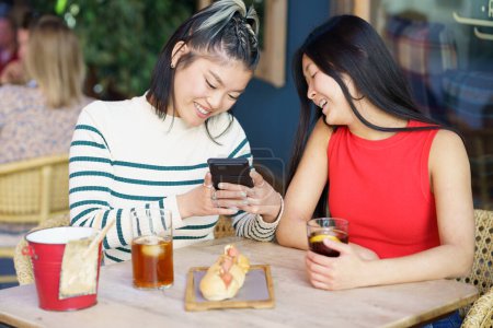 Téléchargez les photos : Contenu jeunes amies asiatiques, en tenues décontractées souriantes et prenant des photos de délicieux repas sur smartphone tout en se rassemblant à la table en bois avec des verres de boissons dans un café - en image libre de droit