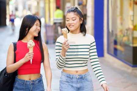 Foto de Feliz joven Asiático mujeres sonriendo y comiendo delicioso helado cono mientras caminan juntos en la calle en la ciudad - Imagen libre de derechos