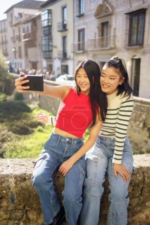 Téléchargez les photos : Heureux jeunes asiatiques amies en vêtements décontractés prenant autoportrait sur smartphone tout en étant assis ensemble et en regardant la caméra à Grenade - en image libre de droit