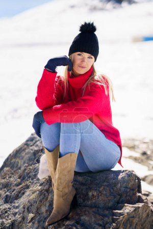 Photo for Blonde young woman sitting on a rock in the snowy mountains in winter, in Sierra Nevada, Granada, Spain. Female wearing winter clothes. - Royalty Free Image