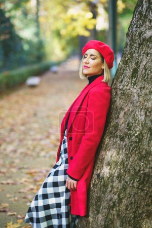 Téléchargez les photos : Vue latérale de la femelle rêveuse aux yeux fermés portant des vêtements de dessus rouges élégants appuyés sur le tronc d'arbre dans le parc d'automne - en image libre de droit