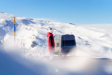 Photo for Anonymous person in red vest cap and gloves looking away while standing on asphalt road, and observing back of moving travel bus against snowy mountain and blue sky - Royalty Free Image