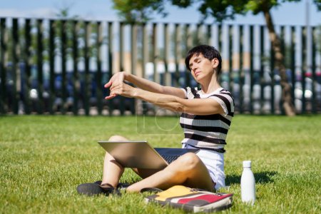 Photo for Female freelancer with laptop and water bottle sitting on lawn while stretching arms in park at sunny day and closed eyes - Royalty Free Image