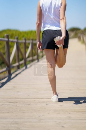 Téléchargez les photos : Femme anonyme en tenue de sport debout sur un pont en bois avec la jambe et le pied tendu vers le haut par derrière, avec la main tout en se réchauffant au soleil contre une campagne floue - en image libre de droit
